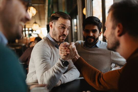 Two men arm wrestling on a wooden table in a pub while their two friends watch closely and cheer them on during a fun night out.
