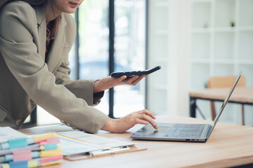 Calculating Efficiency in Modern Office: A focused businesswoman, employing a calculator while immersed in her work, navigates her laptop with precision, exemplifying modern office efficiency.