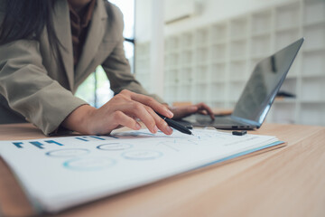 Woman's Focused Analysis: A focused woman, immersed in her work, meticulously examines financial data on a printed document. In the background a laptop rests.