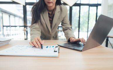 Focused Businesswoman at Her Desk: A sophisticated businesswoman leans over her work, utilizing both a laptop and documents on the desk. Focused, determined, and hard at work.