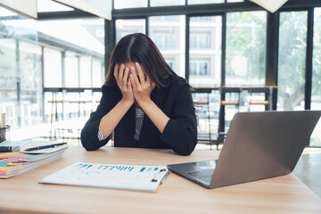 Overwhelmed at Work: A businesswoman, facing challenges, sits at her desk, hands covering her face, with a laptop and documents on the table, reflecting the pressures of the corporate environment.