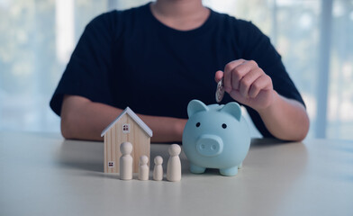 Hand placing a coin into a blue piggy bank beside a wooden house and family figures, representing family saving, financial planning, home budget, security, wealth management, and future stability.