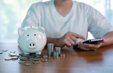 Personal savings and digital money management concept with white piggy bank, stacked coins and person using smartphone on wooden table, representing budgeting, mobile banking.