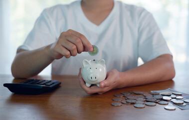 Personal savings and budgeting concept with hand putting coin into white piggy bank on wooden table, calculator and loose coins nearby, representing money management, financial planning.