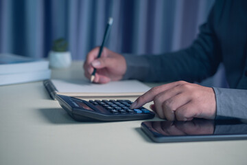 Person calculating budget using calculator and writing notes with pencil at home office desk, personal finance planning, expense tracking, saving money concept, financial management lifestyle.