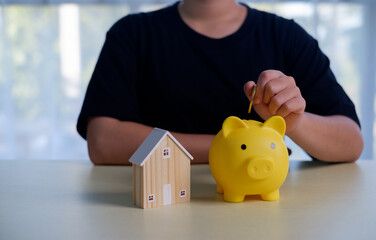 Hand putting a coin into a yellow piggy bank beside a small wooden house, symbolizing saving money for home, financial planning, real estate investment, budgeting, wealth growth, and future security.