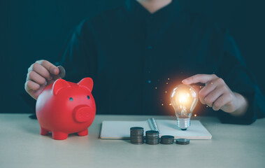 Person putting coin into piggy bank beside glowing light bulb and stacked coins, saving money and financial planning concept, smart investment, creative ideas for personal finance growth.