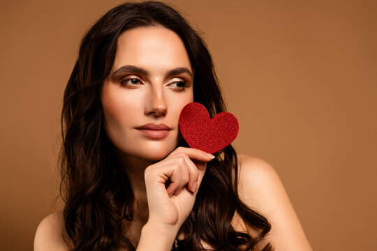 Beautiful brunette woman holding red heart decoration on beige background with elegant makeup and stylish appearance