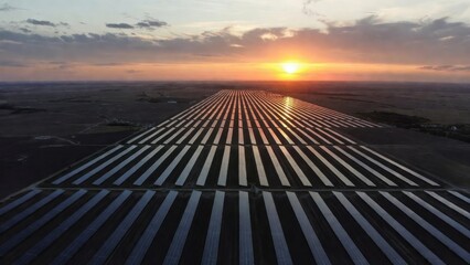 Aerial wide shot of a massive solar panel farm stretching to the horizon at sunset. Rows of photovoltaic panels reflect the golden light of the sun under a picturesque sky, representing renewable ener