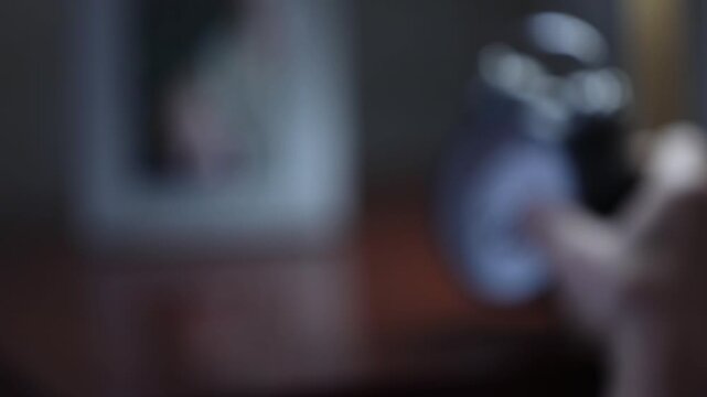 Close-up of wrinkled senior hands holding a classic black alarm clock. The person checks the time and places it on a bedside table in a dimly lit room.

