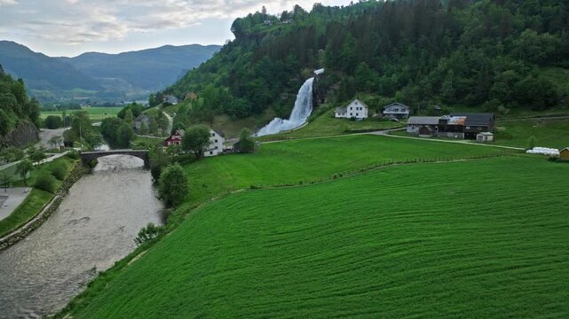 Steinsdalsfossen waterfall above farmland in Norheimsund, Norway. Aerial view combining powerful cascade, green fields, and nearby houses in a summer valley.