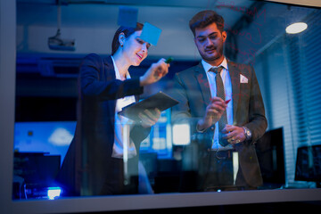 A businessman writing ideas on a glass wall with colorful markers. The woman holds a digital tablet while the man writes Marketing Concept in red.