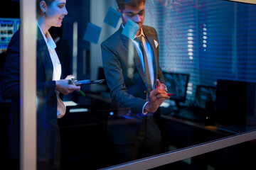 Innovative digital transformation team brainstorming on glass wall. Professional executive writing future business strategy while female colleague analyzes data in modern blue-lit office at night.