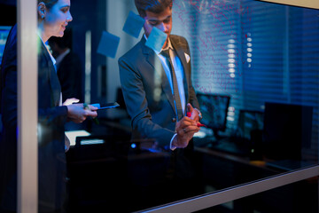 Innovative digital transformation team brainstorming on glass wall. Professional executive writing future business strategy while female colleague analyzes data in modern blue-lit office at night.