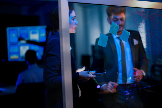 Innovative digital transformation team brainstorming on glass wall. Professional executive writing future business strategy while female colleague analyzes data in modern blue-lit office at night. - Powered by Adobe