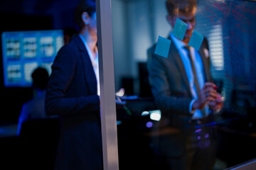 Side view of corporate team brainstorming on glass wall. Businessman writing marketing strategy while female executive holds tablet in modern dark blue office at night.