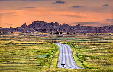 Badlands National Park landscape stands in South Dakota, USA. Scenic view features the winding Loop Road leading towards rugged rock formations and green prairie under a colorful sunset sky