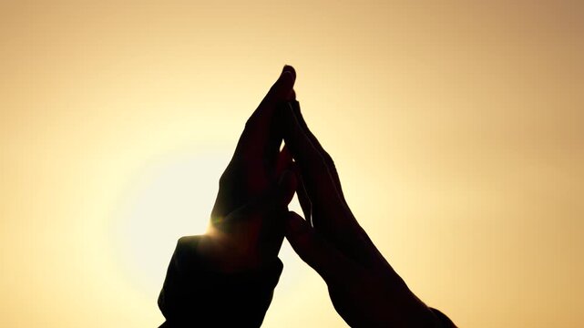 Hands join in prayer silhouette at sunset, palm touch and finger alignment with sun flare between hands, warm golden sky, contemplative unity and connection, peaceful gesture conveying serene hope