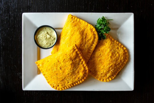 Overhead view of golden baked Jamaican beef patties on a white plate with cilantro sauce on a black table