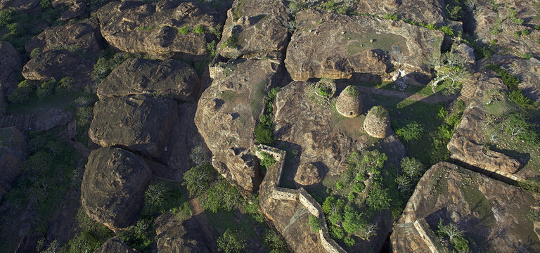 Aerial view of ancient stone structures and walls nestled amidst rugged, sun-kissed boulders and verdant foliage, Badami, Karnataka, India.