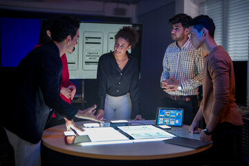 Creative UI/UX design team analyzing mobile app wireframes on illuminated digital touch table. Diverse developers brainstorming web layout strategy in modern high-tech office at night.