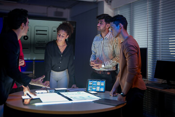 Creative UI/UX design team analyzing mobile app wireframes on illuminated digital touch table. Diverse developers brainstorming web layout strategy in modern high-tech office at night.