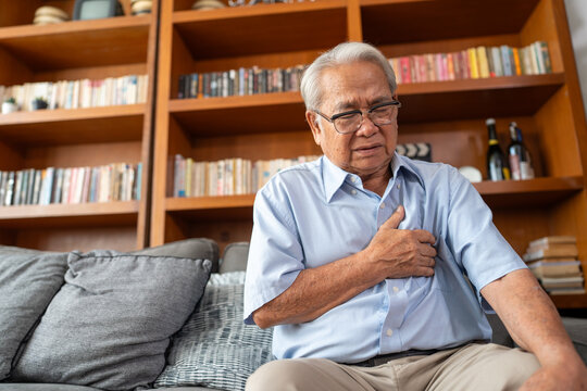 Senior man experiencing chest pain at home. Older man sitting on couch with hand on chest. Heart disease problems.