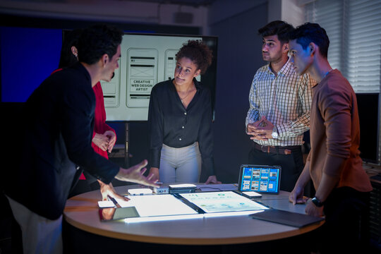 Creative UI/UX design team analyzing mobile app wireframes on illuminated digital touch table. Diverse developers brainstorming web layout strategy in modern high-tech office at night. - Powered by Adobe