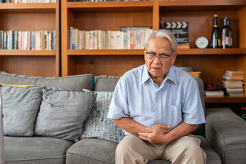 Fototapeta premium Asian elderly man holding stomach pain while sitting on the sofa at home. Senior man experiencing digestive issues or abdominal cramps.