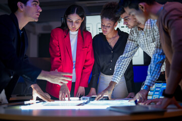 Creative UI/UX design team analyzing mobile app wireframes on illuminated digital touch table. Diverse developers brainstorming web layout strategy in modern high-tech office at night.