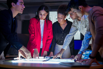 Creative UI/UX design team analyzing mobile app wireframes on illuminated digital touch table. Diverse developers brainstorming web layout strategy in modern high-tech office at night.