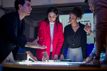 Creative UI/UX design team analyzing mobile app wireframes on illuminated digital touch table. Diverse developers brainstorming web layout strategy in modern high-tech office at night.