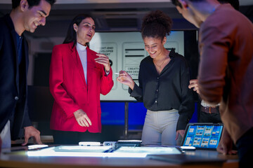 Creative UI/UX design team analyzing mobile app wireframes on illuminated digital touch table. Diverse developers brainstorming web layout strategy in modern high-tech office at night.