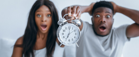 Two people sit on a couch looking shocked as one holds an alarm clock. The light background adds focus to their expressions, showing surprise or disbelief.