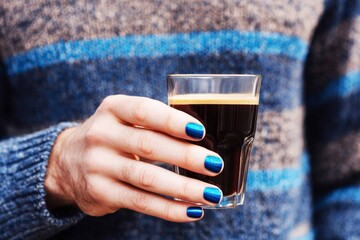 A man sits in a comfortable position, holding a blue coffee cup. His nails are recently manicured in a dark color. He wears a knitted sweater and jeans, creating a casual look