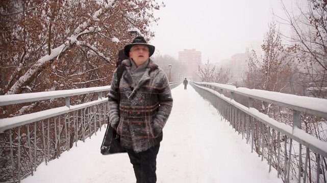 Man in warm clothes with scarf walks along sidewalk among trees and shields himself from heavy snowfall with his hand. Positive mood with smile during extreme winter weather in city.