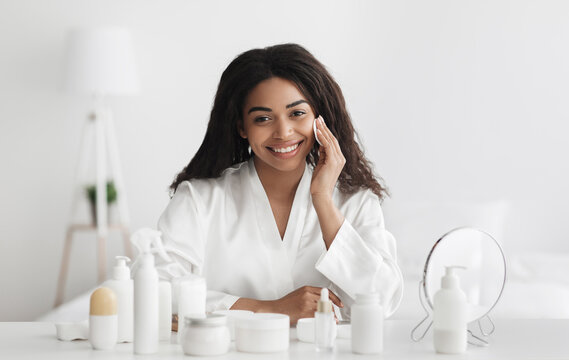 Happy african american woman removing makeup with cotton pad, sitting at dressing table and smiling at camera in bedroom interior. Lady caring for face skin, beauty and facial skincare concept - Powered by Adobe