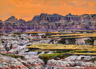 Big Badlands Overlook in Badlands National Park, South Dakota, USA. Scenic view features colorful eroded rock formations and grassy layers under a bright orange sunset sky