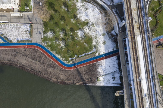 Aerial view of a vibrant red and blue path curving along the stark riverbank contrasting with patches of snow and the adjacent railway bridge, Istanbul, Istanbul, Turkey.
