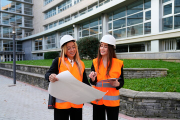 Two young aspiring female architects or engineers wearing safety vests and hard hats analyzing blueprints on location