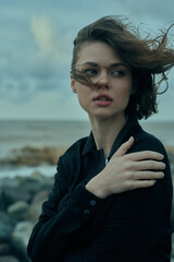 Woman by the sea in windy weather, wearing a dark blouse, dramatic pose, and wind swept hair, capturing moody fashion imagery with coastal scenery and bold contrast, outdoors feeling, no branding,
