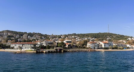 coastal town skyline seen from sea, rows of colorful houses clinging to hillside, waterfront pier and small harbor, calm blue