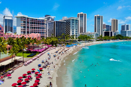 Aerial view of a vibrant beach with turquoise waters meeting the sandy shore, backed by tall buildings under a clear blue sky, Honolulu, Hawaii, United States.