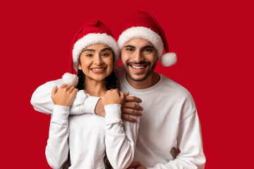 A cheerful couple wears Santa hats, embracing each other in a cozy pose. The bright red background enhances their joyful expression, capturing the spirit of the holiday season.