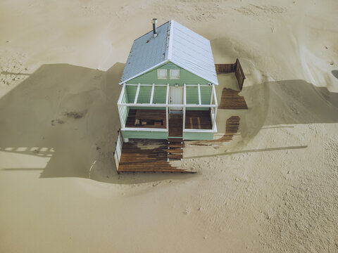 Aerial view of a lone, mint-green house with weathered wooden accents stands nestled amidst a vast expanse of beige sand, contrasting textures under a muted sky, Costa da Caparica, Setubal, Portugal.