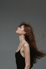 Fototapeta premium Profile of a young woman with long hair flowing, wearing a black dress, exuding calm and grace against a neutral gray background.