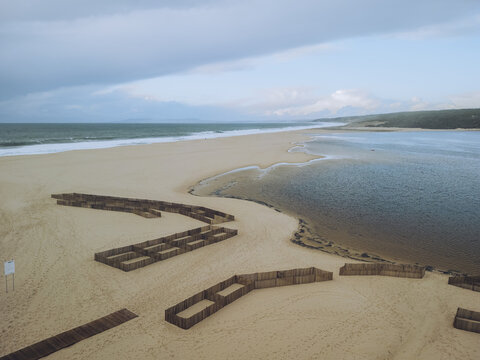 Aerial view of the tranquil lagoon meeting the vast ocean under a serene sky, framed by sandy shores and constructed barriers, Lagoa de Albufeira, Setubal, Portugal.