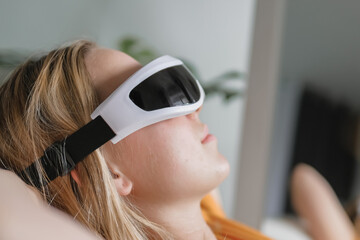 Gen Z woman unwinds  home smart eye massagerЧwellness tech  recovery, mindfulness and stress relief. Cozy greenery and soft light set a self-care, digital-detox mood, promoting balance, calm wellbeing
