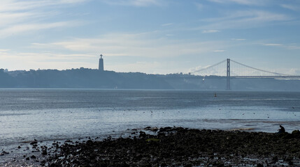 A view of the 25 de Abril Bridge and Cristo Rei statue in Lisbon, Portugal at sunset with sailboats on the river.
