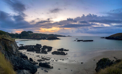Wide coastal view at dusk with calm sea, rocky shore, sandy beach, and dramatic clouds reflecting soft light, creating a peaceful natural landscape